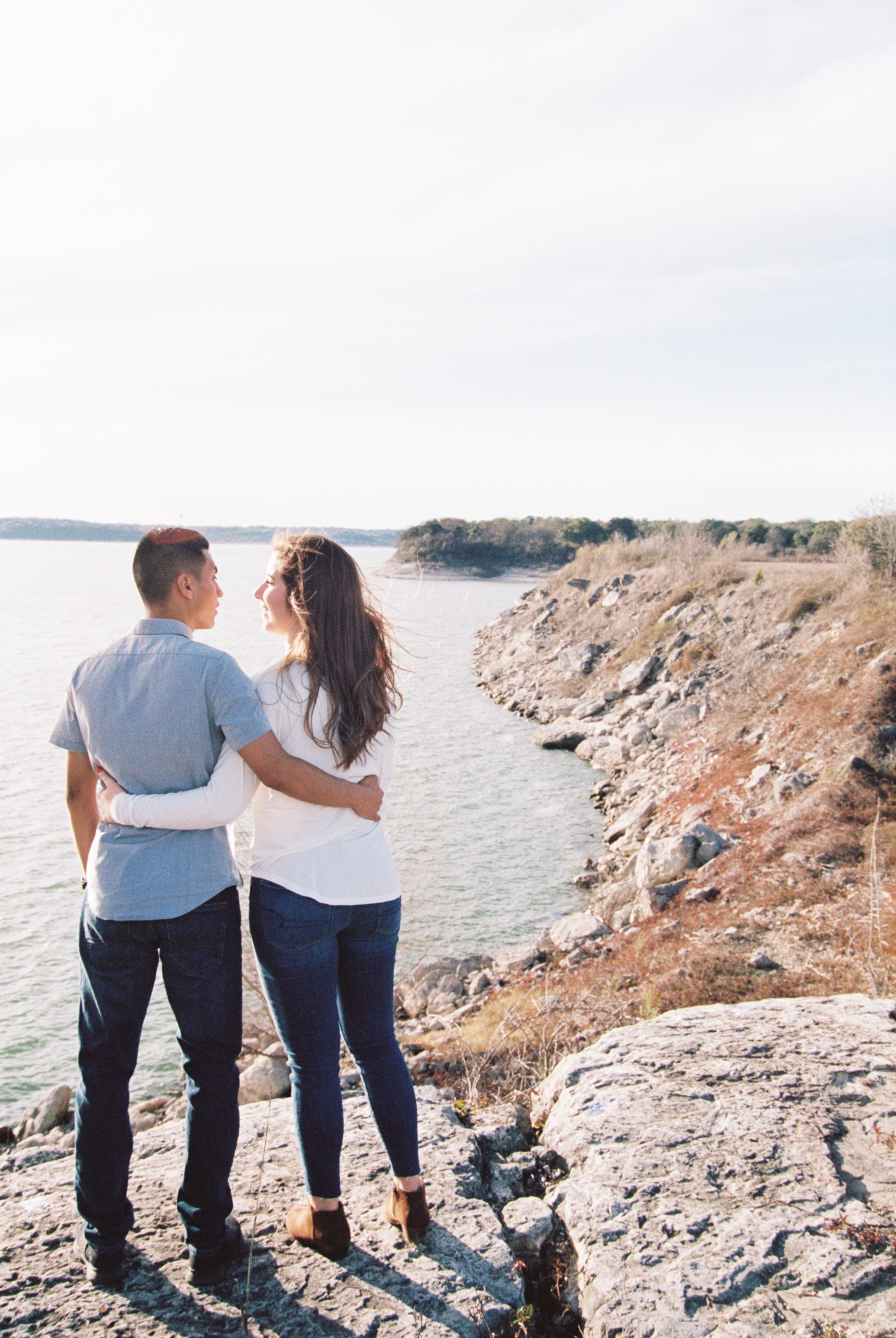 a couple looks at each other while on the edge of a cliff over looking the water