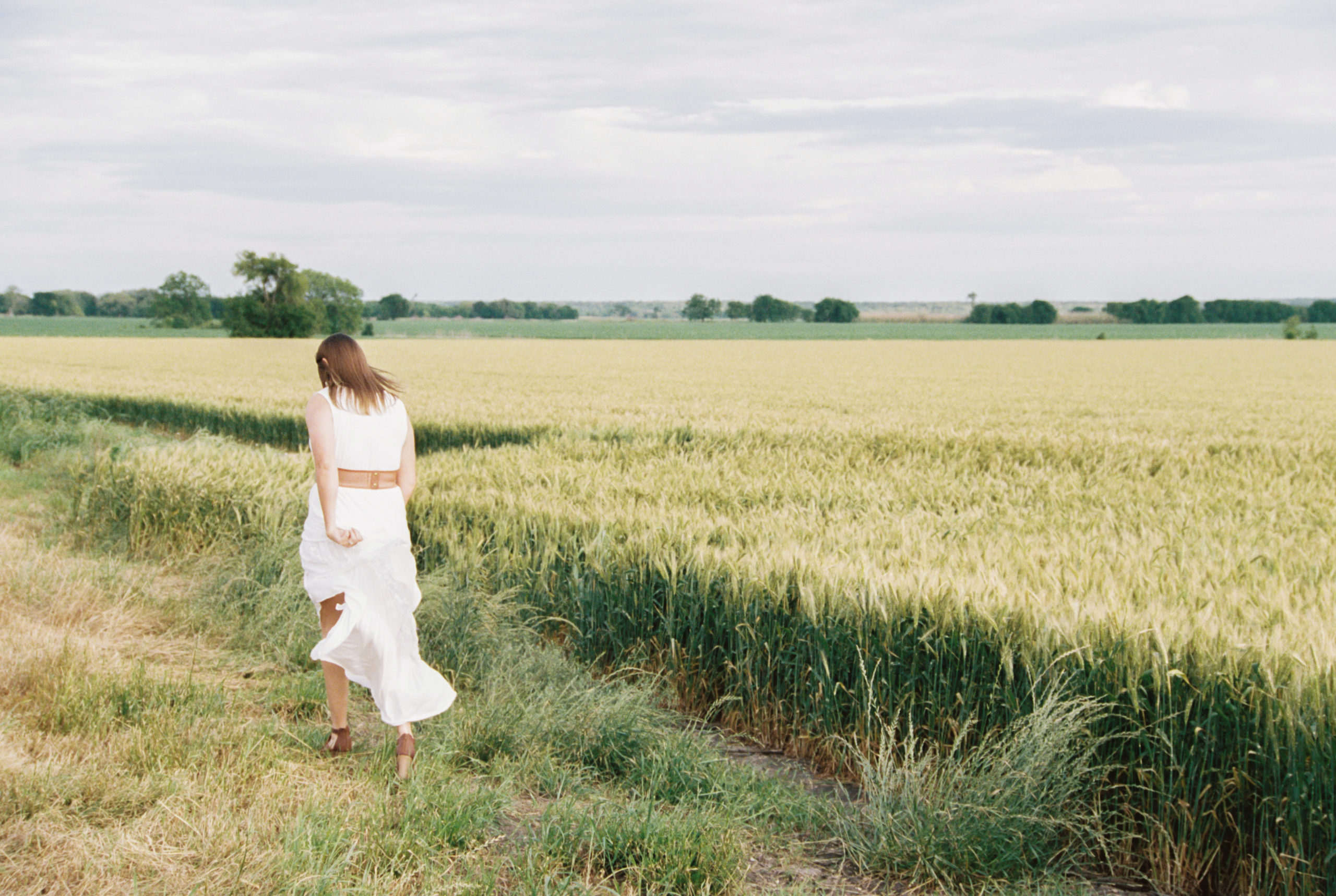 A woman in a white dress walks off in the distance, next to the wheat field swing the bottom of her dress