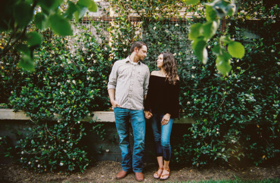 a man and a woman stand in front of a honeysuckle wall holding hands and looking at each other.