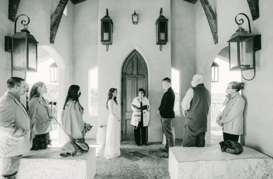 A black and white image of elle and henry standing across from each other at Chapel Dulcinea as other watch them get married.