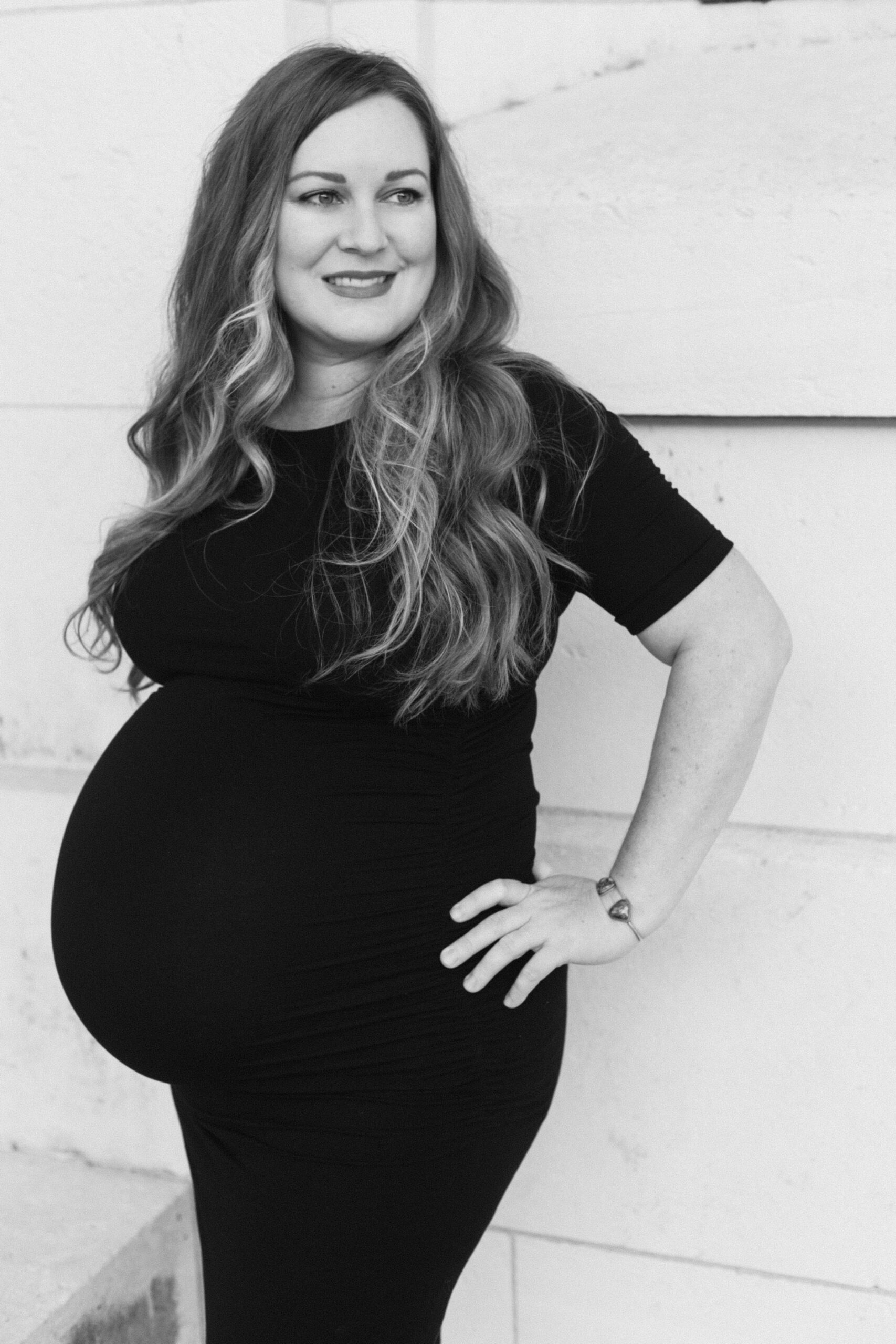 Black and white image of a pregnant woman in a black dress with a hand on her hip looking past the camera. This image is being used as a feature on the Rae Allen Investment Page.