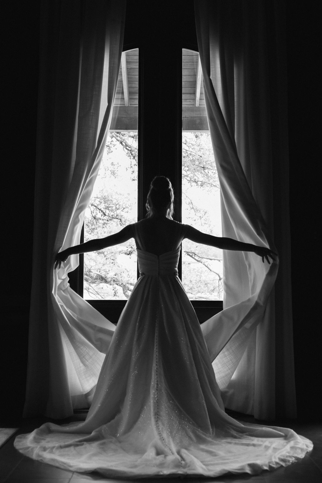 This is a black and white image of the back on the bride, while she is throwing the curtains aside in the bridal suite. Shot by Rae Allen Photography