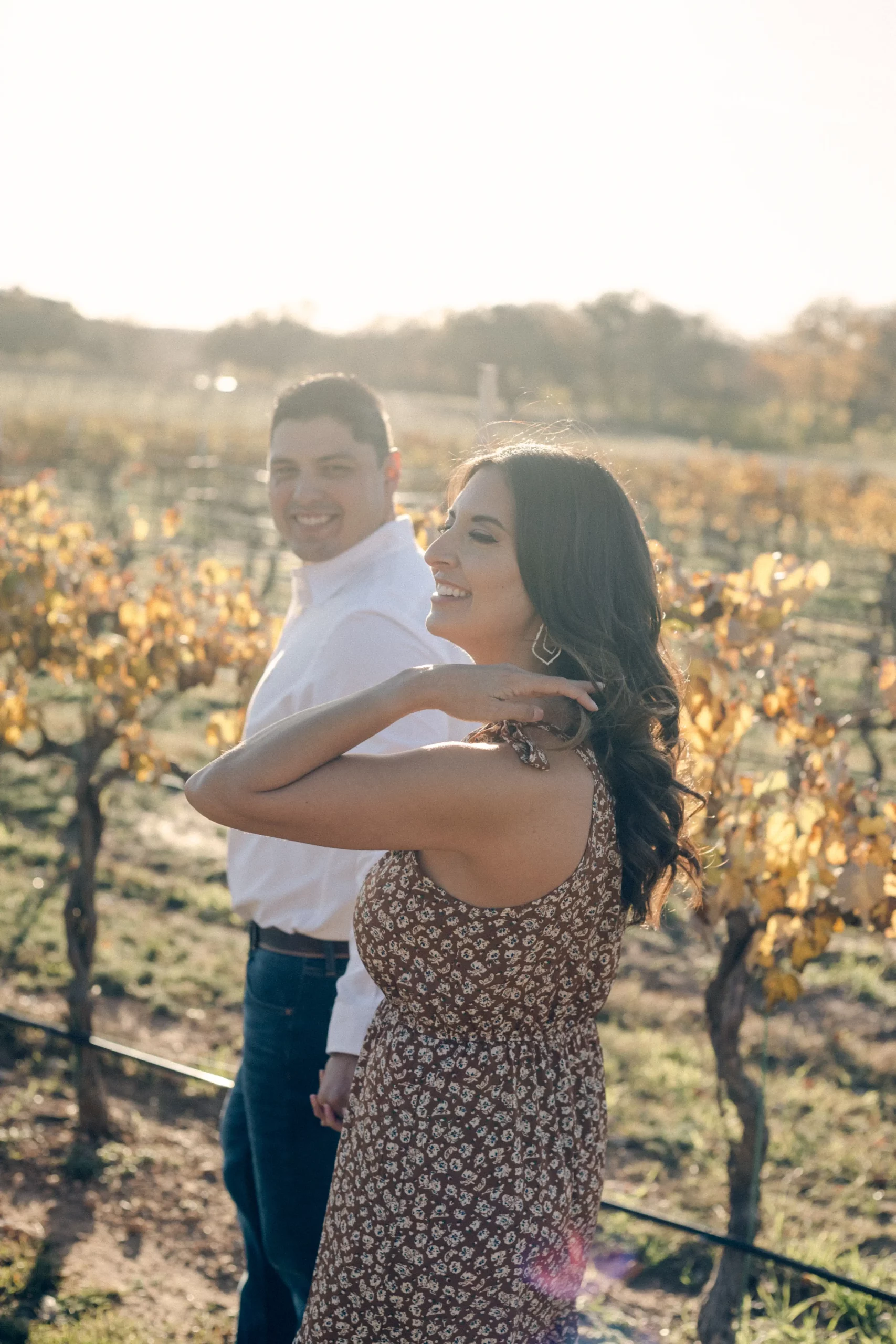 Rae Allen Photography| Janessa brushes back her hair as David looks at her during their engagement shoot at Fiesta Winery