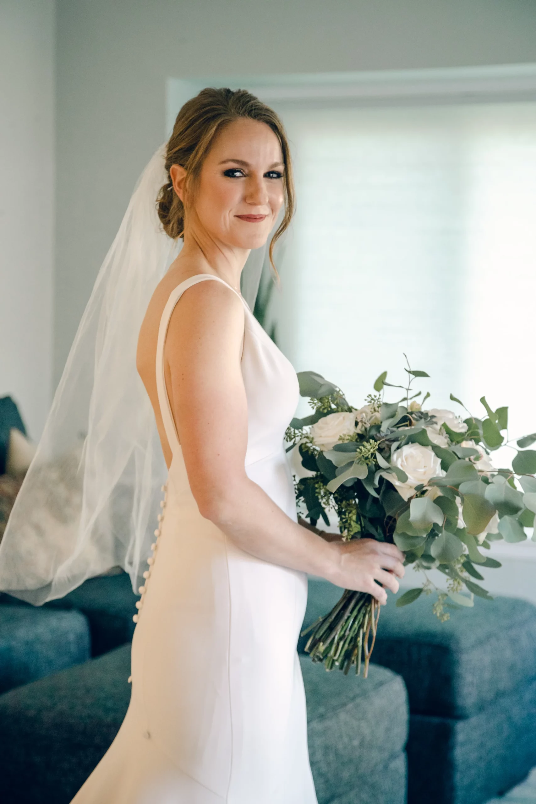 Megan takes a bridal portrait with Rae Allen Photography in her living room hold her white rose and greenery bouquet